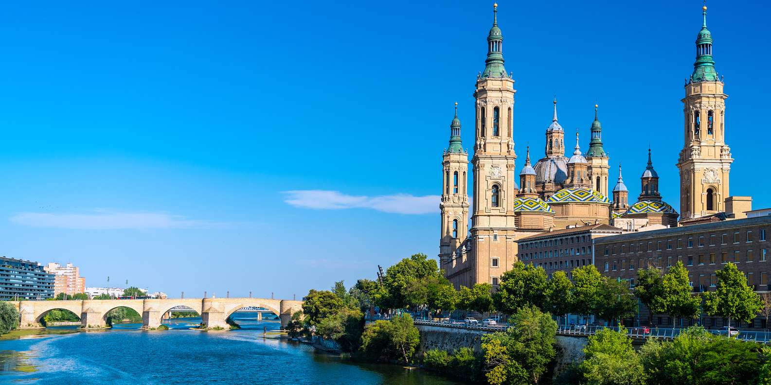 View of the Basilica of Our Lady of the Pillar in Zaragoza, with its iconic colorful tiled domes and tall towers on the banks of the Ebro River, next to the historic Stone Bridge under a clear sky.