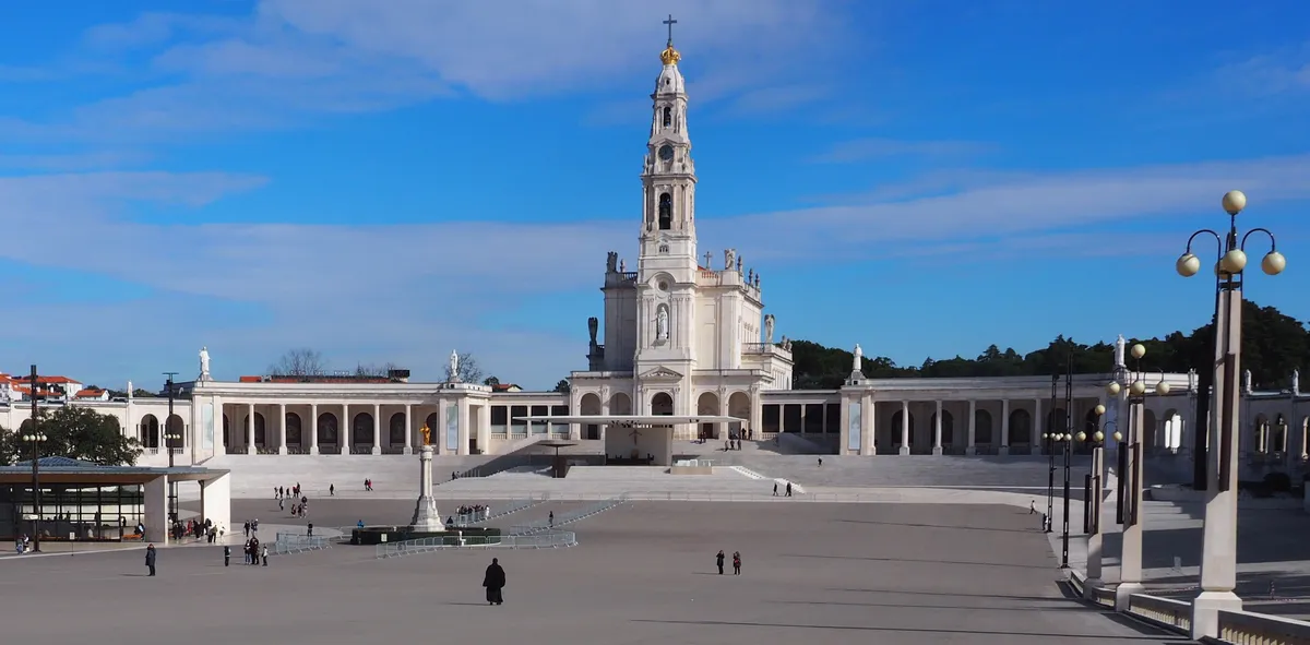 White facade of the Basilica of Fatima with its tall central tower and side colonnades surrounding the prayer square.